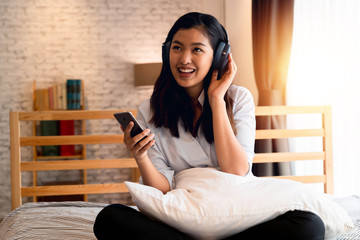 Obraz premium Happy young Asian woman in earphones singing and listening to music while sitting on bed and holding mobile phone at home.
