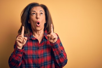 Middle age beautiful woman wearing casual shirt standing over isolated yellow background amazed and surprised looking up and pointing with fingers and raised arms.