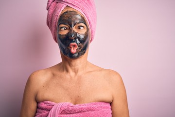 Middle age brunette woman wearing beauty black face mask over isolated pink background making fish face with lips, crazy and comical gesture. Funny expression.