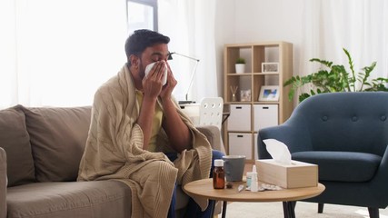 healthcare, cold, hygiene and people concept - sick indian man in blanket sneezing and blowing nose with paper tissue at home