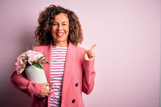 Middle age beautiful woman holding vase with flowers over isolated pink background very happy pointing with hand and finger to the side