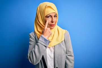 Middle age brunette business woman wearing muslim traditional hijab over blue background Pointing to the eye watching you gesture, suspicious expression