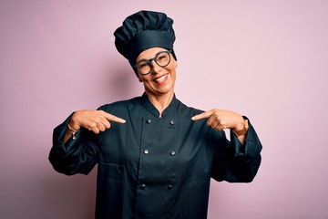 Middle age brunette chef woman wearing cooker uniform and hat over isolated pink background looking confident with smile on face, pointing oneself with fingers proud and happy.