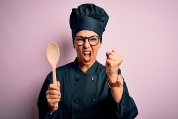 Middle age brunette chef woman wearing cooker uniform and hat holding wooden spoon annoyed and...