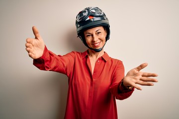 Middle age motorcyclist woman wearing motorcycle helmet over isolated white background looking at the camera smiling with open arms for hug. Cheerful expression embracing happiness.