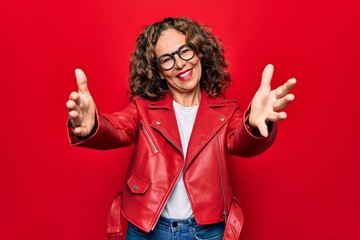 Middle age beautiful woman wearing casual red jacket and glasses over isolated background looking at the camera smiling with open arms for hug. Cheerful expression embracing happiness. - obrazy, fototapety, plakaty
