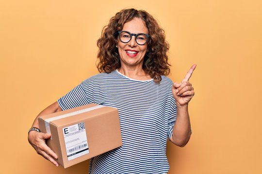 Middle age beautiful woman holding cardboard package with label over yellow background smiling happy pointing with hand and finger to the side