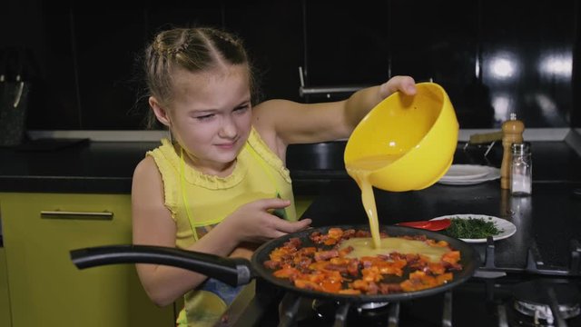 Smart girl learning to cook. Happy and serious young mistress children to cook a Neapolitan egg fried omelette from sausage. Modern Built In Kitchen Appliances in green black color.