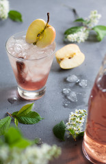 Refreshing drink with ice from apples with cinnamon in a glass on a gray table with scattered flowers and leaves