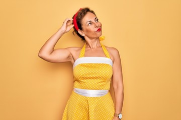 Middle age senior pin up woman wearing 50s style retro dress over yellow background confuse and wondering about question. Uncertain with doubt, thinking with hand on head. Pensive concept.