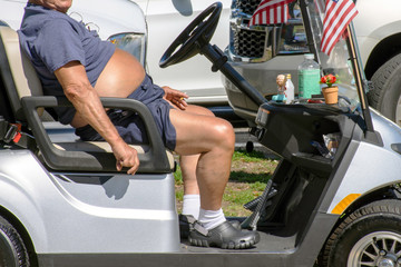 Overweight and out of shape, a senior man with a beer belly smokes a cigarette while riding a golf...