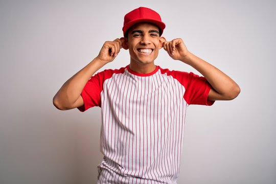 Young handsome african american sportsman wearing striped baseball t-shirt and cap Smiling pulling ears with fingers, funny gesture. Audition problem