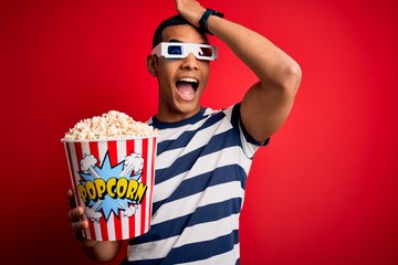 Young handsome african american man watching movie using 3d glasses eating popcorns surprised with...