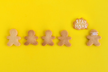 Homemade cookies in shapes of people with inscription ‘Stay home’ and with face medical mask on yellow background, top view. Sweet shortbread with white glaze. Social distancing concept.