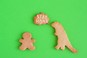 Homemade cookies in shapes of dinosaur and man with inscription ‘Stay home’ on green background, top view. Sweet shortbread with white glaze. Social distancing concept.