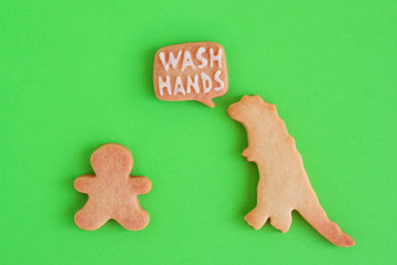Homemade cookies in shapes of dinosaur and man with inscription ‘Wash hands’ on green background, top view. Sweet shortbread with white glaze. Social distancing concept.