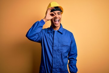 Young handsome african american worker man wearing blue uniform and security helmet doing ok gesture with hand smiling, eye looking through fingers with happy face.
