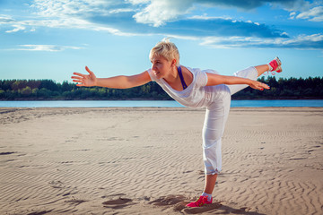 Blond-haired woman in a white tracksuit on a sandy beach performs swinging legs and arms