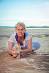 A fair-skinned blonde in a white tracksuit sits on a sandy beach and does stretching exercises, stretches her arms to the feet