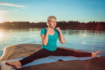 A blonde woman with a short haircut on a sandy beach near the water sits on a blue rug and exercises