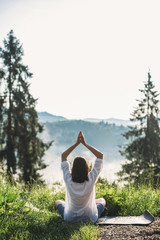 Young woman practicing yoga and sitting among grass in sunny light on background of morning mountains. Meditation. Calm tranquil moment, connection with nature. Copy space