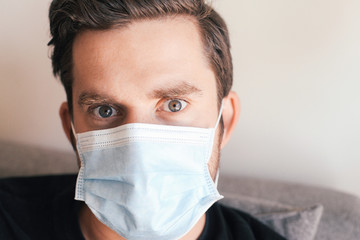 Close up portrait of a man in medical mask, green grey eyes, looking worried, sitting on a couch.