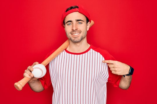 Young handsome sporty man with blue eyes playing baseball wearing cap holding bat and ball with surprise face pointing finger to himself