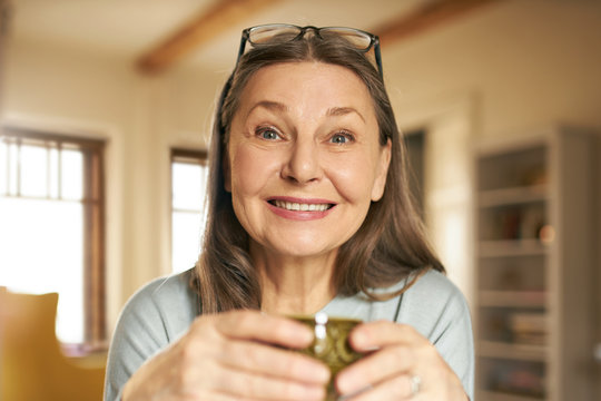 Close up image of charming female pensioner with glasses on head smiling, enjoying fresh coffee in the morning. Energetic cheerful middle aged woman in casual clothes holding cup, drinking tea at home