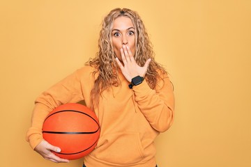 Beautiful blonde sportswoman doing sport holding basketball ball over yellow background covering mouth with hand, shocked and afraid for mistake. Surprised expression