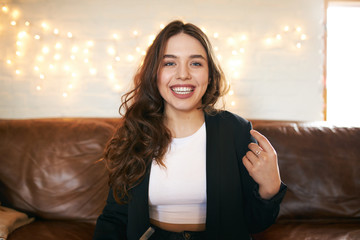 Cute adorable teenage girl sitting on leather couch in living room looking with broad happy smile,...