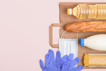 Essential food set. Craft package, midical mask and gloves, bottles of oil and milk, spaghetti and baguette on the light pink background. Safe food delivery. Copy space on the left. Top view.