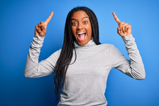 Young african american woman standing wearing casual turtleneck over blue isolated background smiling amazed and surprised and pointing up with fingers and raised arms.