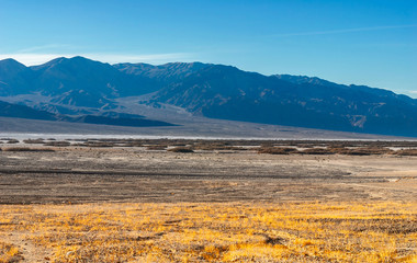 A stony valley is overgrown with rare dry grass against a mountain range.