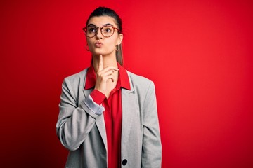 Young beautiful brunette businesswoman wearing jacket and glasses over red background Thinking concentrated about doubt with finger on chin and looking up wondering