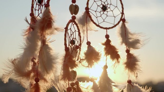 Dreamcatcher with orange feathers, swaying with light wind slow motion slide agains blue sky at sunset in summer. Amulet boho style, Indian style. Bright disk of the sun, the rays of the sun