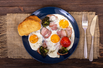 Tasty breakfast with fried eggs, bacon, fried broccoli and tomato, toasts in the plate that is on textured burlap, on wood background, top view