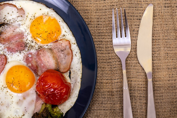 Breakfast with fried eggs, pork ham, broccoli and tomato in a plate that is on textured burlap, fork and knife. Macro photography