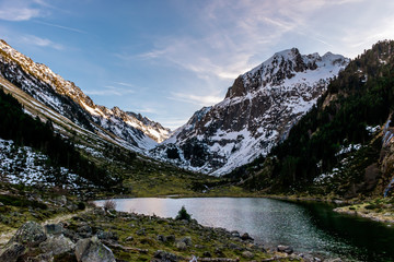 Naklejka na ściany i meble A picturesque landscape view of the high snow capped Pyrenees mountains on a winter evening with a hiking footpath and a lake with still water (Lac de Suyen, Arrens-Marsous, Hautes-Pyrenees, France)