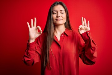 Young beautiful woman with blue eyes wearing casual shirt standing over red background relax and smiling with eyes closed doing meditation gesture with fingers. Yoga concept.