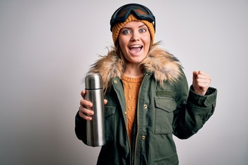 Young beautiful skier woman with blue eyes wearing ski goggles drinking thermo with coffee screaming proud and celebrating victory and success very excited, cheering emotion