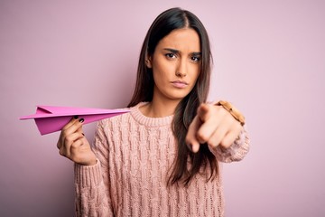 Young beautiful brunette woman holding paper airplane over isolated pink background pointing with finger to the camera and to you, hand sign, positive and confident gesture from the front