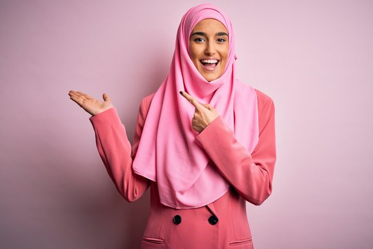 Young beautiful brunette businesswoman wearing pink muslim hijab and business jacket amazed and smiling to the camera while presenting with hand and pointing with finger.