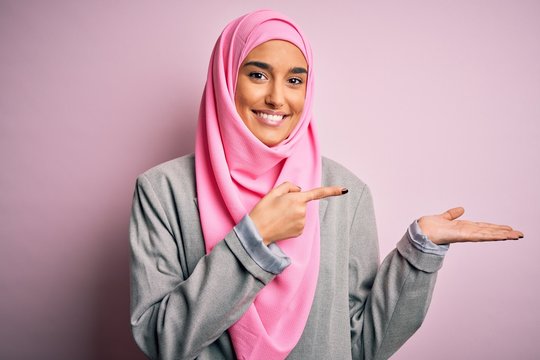 Young beautiful brunette businesswoman wearing pink muslim hijab and business jacket amazed and smiling to the camera while presenting with hand and pointing with finger.