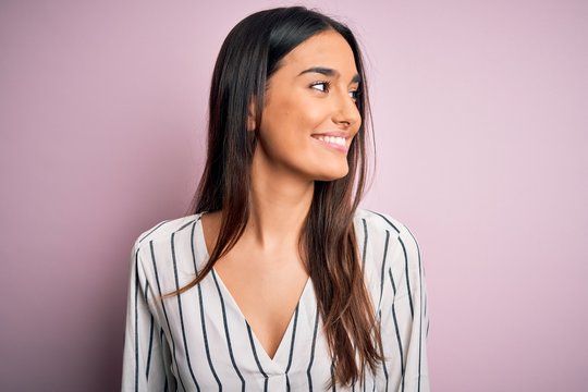 Young beautiful brunette woman wearing casual striped shirt over isolated pink background looking away to side with smile on face, natural expression. Laughing confident.