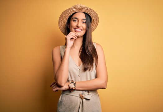 Young beautiful brunette woman on vacation wearing casual dress and hat looking confident at the camera with smile with crossed arms and hand raised on chin. Thinking positive.
