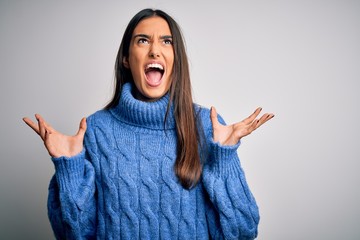 Young beautiful brunette woman wearing casual turtleneck sweater over white background crazy and mad shouting and yelling with aggressive expression and arms raised. Frustration concept.