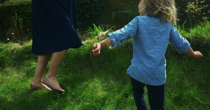 Little preschooler chasing bubbles in garden with his mother