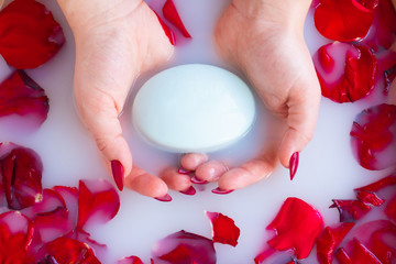 Top view female hands hold soap against the background of milk or white water with rose petals