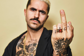 Young aggressive provocative male in black coat over naked tattooed torso showing middle finger gesture while standing against beige background