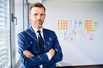 Portrait of confident adult businessman standing at office with crossed arms, looking at camera